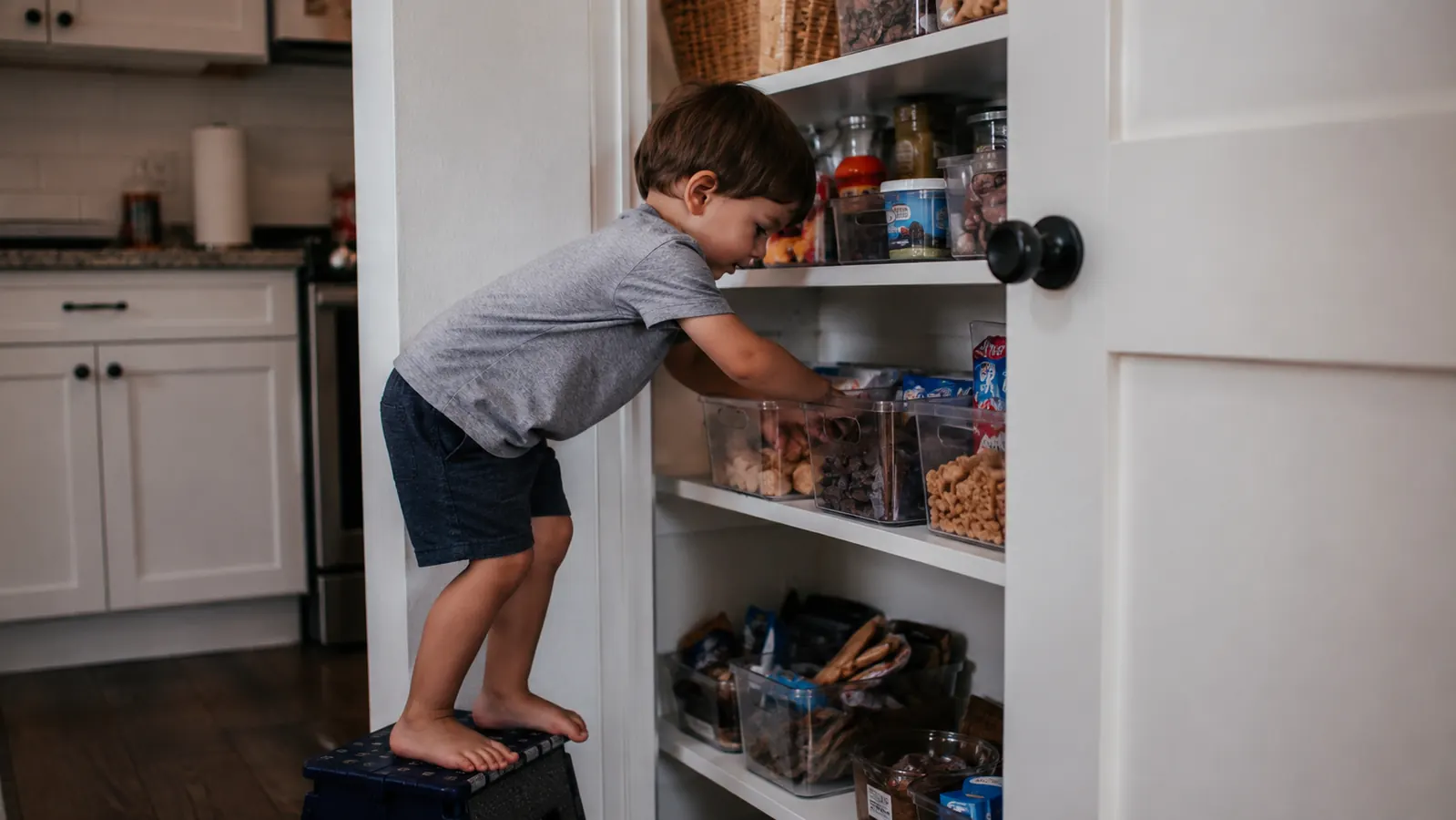Child reaching for a snack inside a clear labeled pantry bin on a low shelf