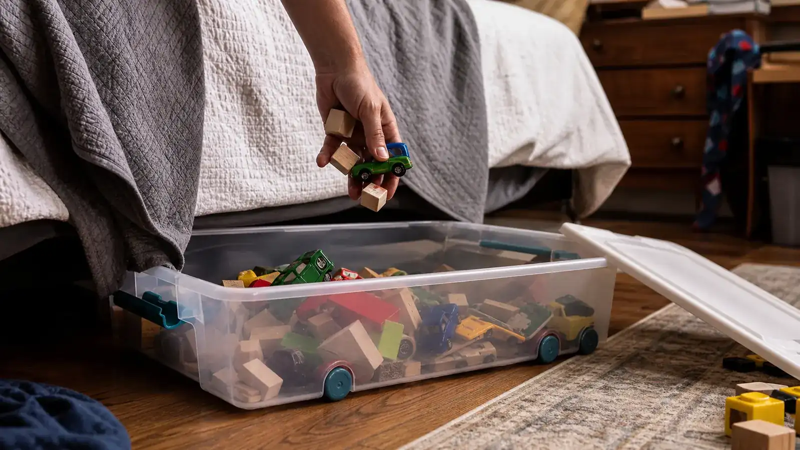 A hand sorting small toy cars and action figures into an open clear plastic storage bin on a bedroom floor